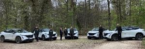 Bild mit fünf weiß-schwarzen E-Autos und sieben Personen in einem grünen Wald 