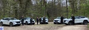 Bild mit fünf weiß-schwarzen E-Autos und sieben Personen in einem grünen Wald 